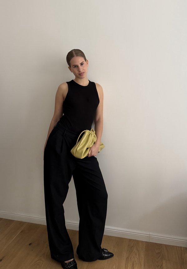 Czarna bezrękawnikowa bluzka, czarne spodnie o wysokim stanie, trzymająca miękką, żółtą skórzaną torebkę. Gładkie materiały i luźny, szeroki krój.