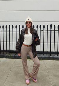 Corta de couro castanho, gorro de malha creme, suéter de gola alta branco com nervuras, calças beiges de boca de sino, sapatos plataforma rosa com riscas pretas; segurando uma chávena de café preto.