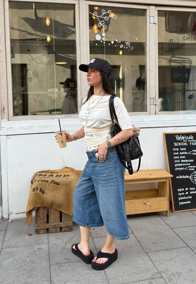 Conjunto casual que incluye una camiseta blanca de manga corta, culottes de mezclilla de pierna ancha, sandalias de plataforma negras y una mochila negra; sostiene un café helado.