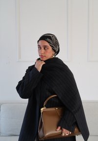 Manteau rayé foncé à surface texturée, drap é sur les épaules. Contient un sac à main marron moyen structuré avec un fermoir doré. Canapé ivoire en arrière-plan.