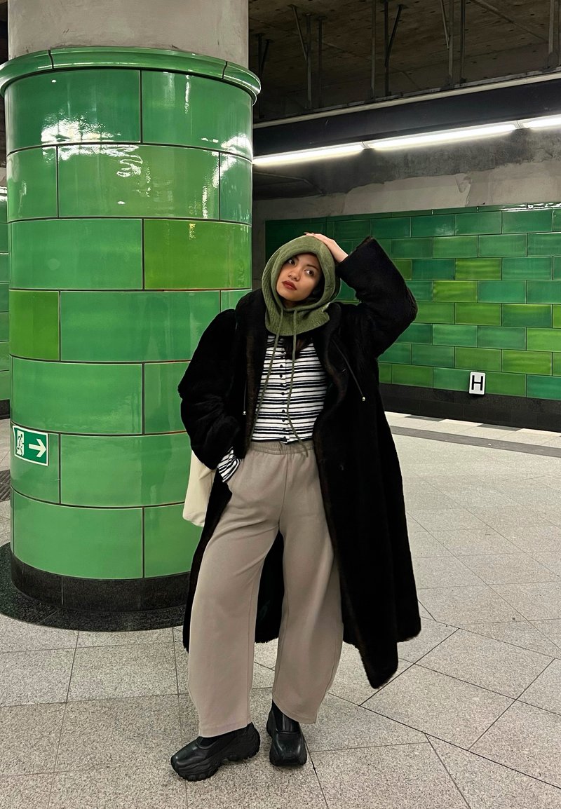 Jeune personne en manteau noir et sweat à capuche vert debout dans une station de métro carrelée aux murs verts et avec un grand pilier.