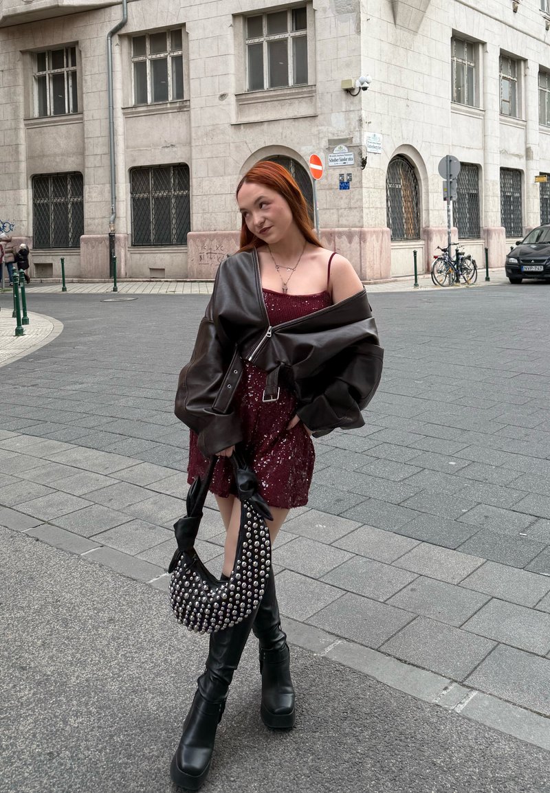 Robe à sequins bordeaux brillante, veste en cuir noire oversize, bottes à plateforme noires montant jusqu'aux genoux, et sac à main clouté. Scène urbaine en rue.