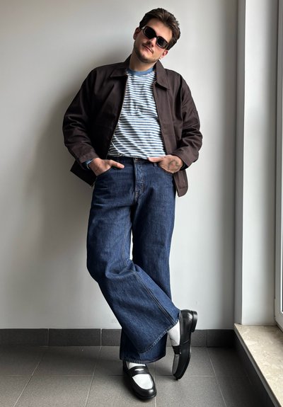 El modelo lleva una chaqueta marrón sobre una camiseta de rayas azules, combinada con unos vaqueros de pierna ancha de mezclilla azul y zapatos negros con detalles blancos.