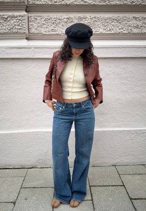 Mujer con gorra negra, chaqueta de cuero marrón, top crema con botones, vaqueros de pierna ancha y zapatos beige con puntera partida, apoyada contra una pared texturizada mirando hacia abajo.