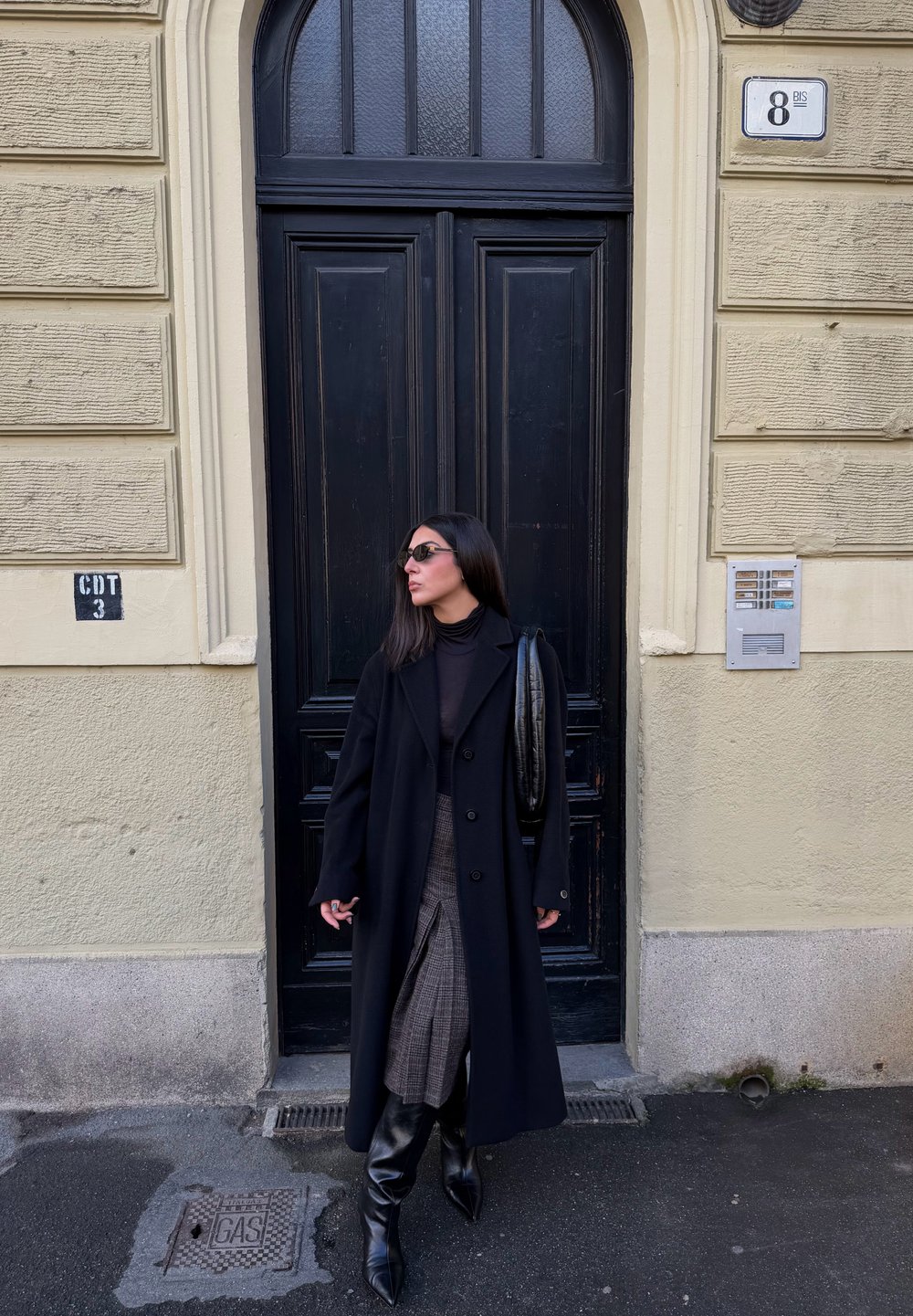 Long manteau noir, haut noir ajusté, jupe à carreaux, bottes noires montant jusqu'aux genoux, lunettes de soleil rondes ; debout près d'une porte noire et d'un mur texturé.