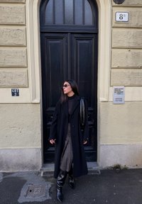 Long manteau noir, haut noir ajusté, jupe à carreaux, bottes noires montant jusqu'aux genoux, lunettes de soleil rondes ; debout près d'une porte noire et d'un mur texturé.