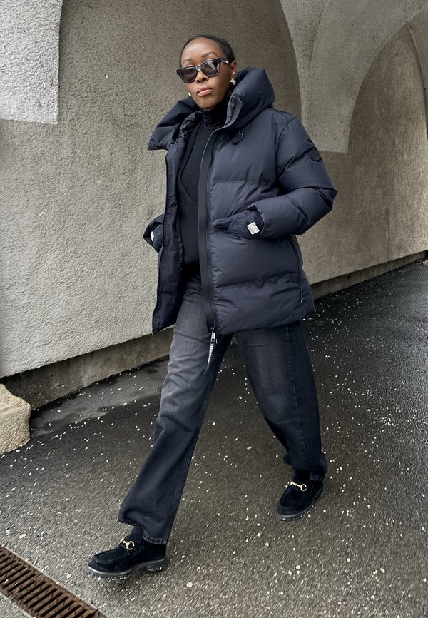 Personne marchant en plein air portant une doudoune noire, un col roulé noir, un jean large gris, des mocassins noirs, des lunettes de soleil foncées et des gants gris.