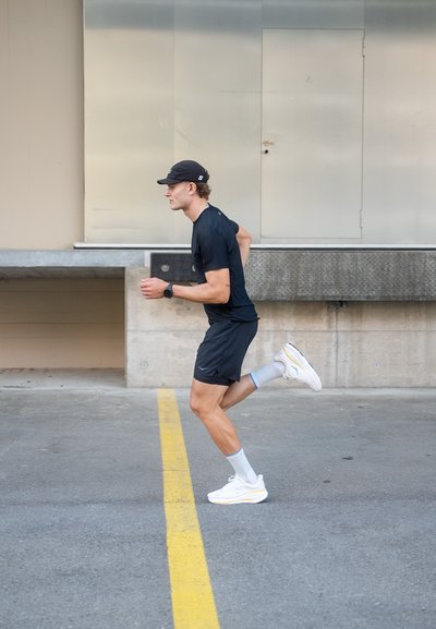 Corredor masculino atlético con camiseta y pantalones cortos negros, que lleva una gorra negra y zapatillas blancas con detalles amarillos, corriendo sobre una superficie gris.