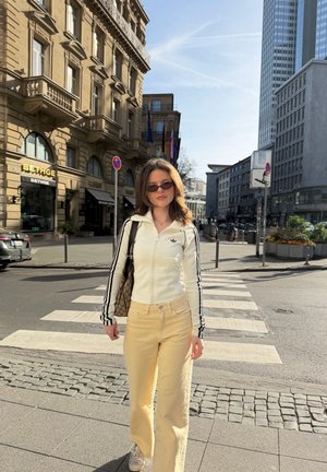 Joven mujer con gafas de sol, chaqueta blanca y pantalones amarillos caminando por un paso de peatones en la ciudad con edificios históricos y modernos al fondo.
