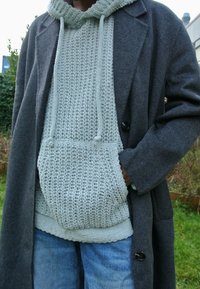 Cappotto grigio in lana su una felpa a maglia verde chiaro con un motivo testurizzato e cordini, abbinato a jeans in denim blu.