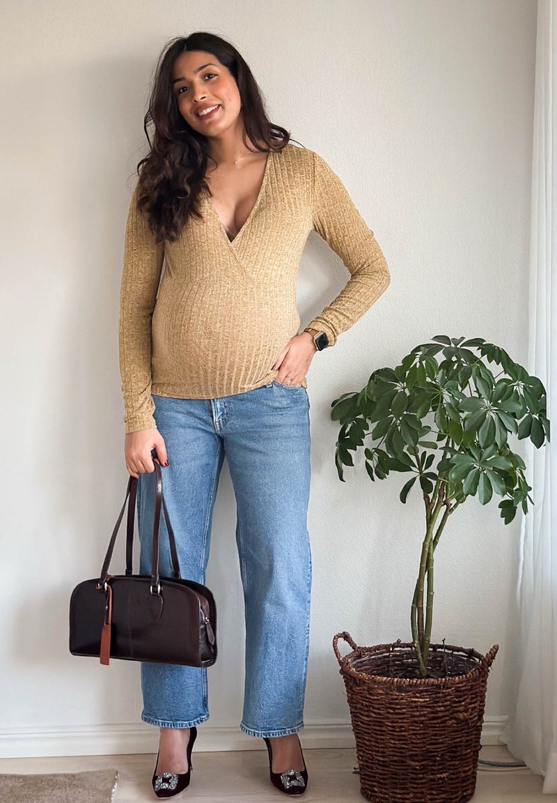 Femme enceinte souriante à l'intérieur, portant un haut beige à manches longues, un jean bleu, des talons noirs, tenant un sac à main marron foncé, debout près d'une plante en pot.