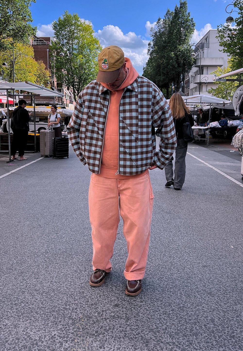 Giacca a scacchi marrone e blu, felpa corallo e pantaloni corallo. Scarpe marroni con dettagli a catena. Ambientazione di mercato all'aperto con bancarelle.