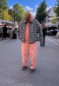 Giacca a scacchi marrone e blu, felpa corallo e pantaloni corallo. Scarpe marroni con dettagli a catena. Ambientazione di mercato all'aperto con bancarelle.