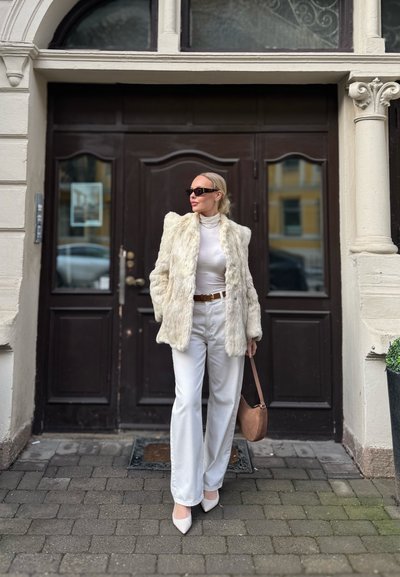 Mujer vestida con abrigo de piel blanco, suéter de cuello alto blanco, pantalones blancos, tacones blancos, gafas de sol y lleva un bolso marrón claro, de pie en una calle empedrada.
