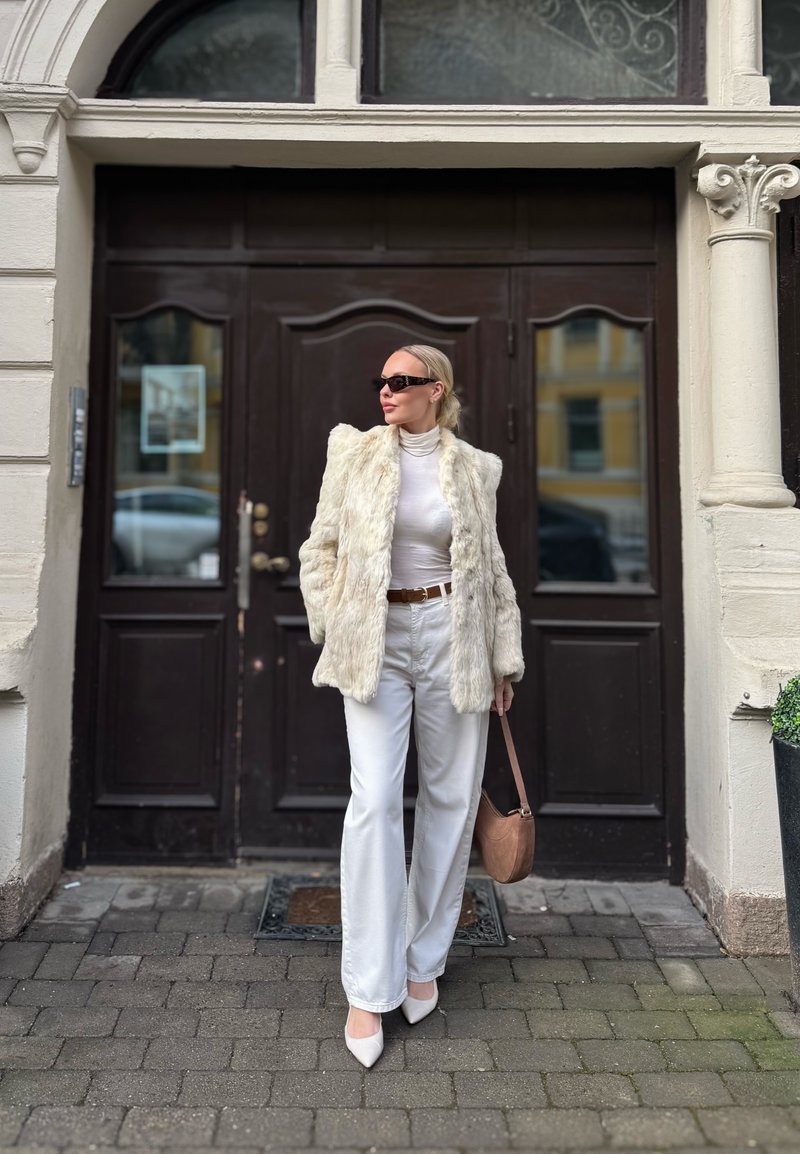 Femme portant un manteau en fourrure blanc, un col roulé blanc, un pantalon blanc, des talons blancs, des lunettes de soleil, et tenant un sac à main beige, debout dans une rue pavée.