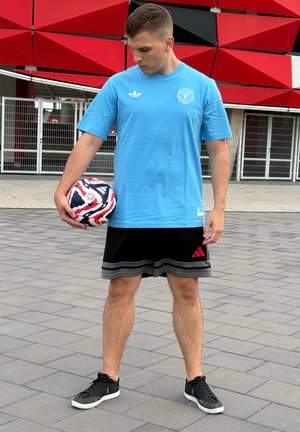 Camiseta de algodón azul claro de manga corta, pantalones cortos negros con el logo rojo de Adidas, sosteniendo un balón de fútbol con estampado, de pie sobre una superficie de baldosas.
