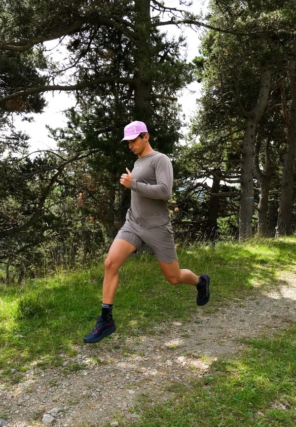 Homme en tenue de sport grise et casquette rose courant sur un sentier forestier entouré de grands arbres et d'herbe verte.