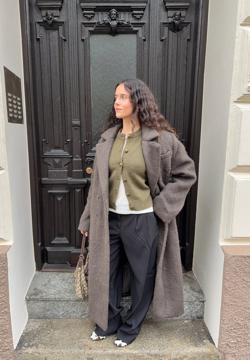 Casaco de lã castanho oversize, cardigan verde-oliva, camisa branca, calças pretas de perna larga, sapatos com estampa de vaca, mala, em pé junto a uma porta de madeira.