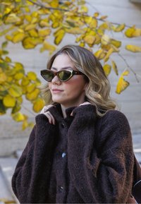 Pruun ülikare pehme mantel mustade nuppudega, kombineeritud roheliste lõhestatud kassisilmaga päikseprillidega, millel on kilpkonna kestast raam, taustal kollased lehed.