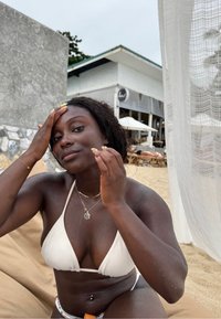 Vit triangelformad bikiniöverdel, flerfärgad midja, slät mörk hy med vattendroppar. Hon håller tillbaka sitt hår och sitter på en sandstrand.