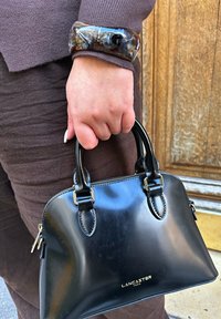 Melna ādas rokassoma ar apaļu formu, diviem augšējiem rokturiem, zelta krāsas detaļām un logotipu. Valdīta ar biezas, tumšas krāsas aproci.