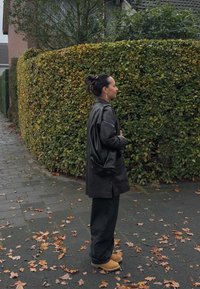 Μαύρο δερμάτινο Oversized μπουφάν, σκούρο παντελόνι και καφέ ξύλινες παντόφλες. Στηρίζει τον εαυτό του δίπλα σε ένα καλοκουρεμένο θάμνο σε έναν πεζόδρομο γεμάτο φύλλα.