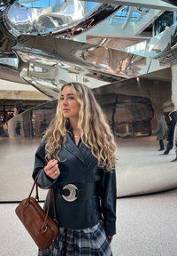 Jeune femme aux longs cheveux bouclés, portant une veste en cuir noire et une jupe à carreaux, tenant un sac à main marron, debout près d'une sculpture en métal brillant.