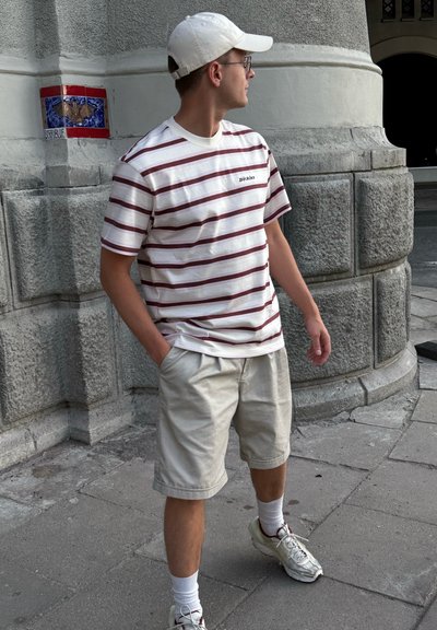 Camiseta blanca a rayas con rayas burdeos y logo, pantalones cortos crema, calcetines blancos y zapatillas grises en un entorno urbano.