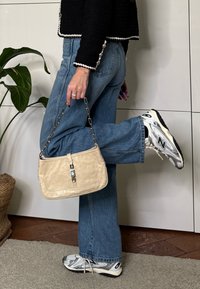 Personne portant un jean bleu, une veste noire et des baskets New Balance blanches, tenant un sac à main beige avec une bandoulière chaîne, une jambe levée vers l'arrière.