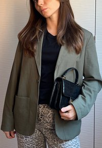 Blazer verde-oliva com um único botão e bolso, combinado com uma t-shirt preta e calças de estampado de leopardo. A modelo segura uma mala preta texturizada.