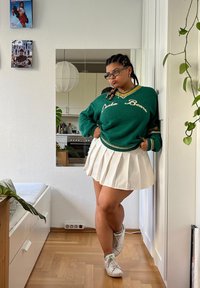 Πράσινο πουλόβερ με κείμενο "Drake Bears", λευκή πλισέ φούστα και λευκά αθλητικά παπούτσια. Ο χώρος έχει ξύλινο δάπεδο και εσωτερικά φυτά.