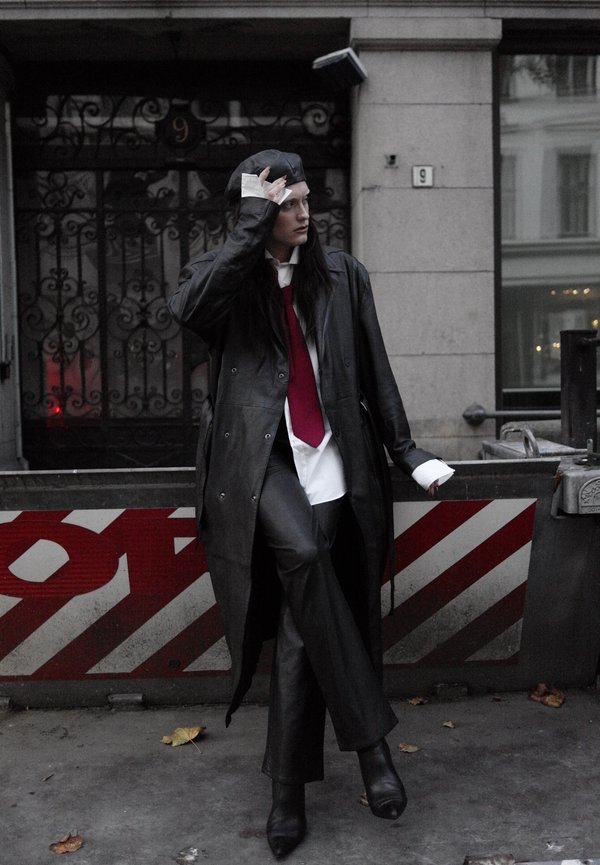 Una persona con un abrigo negro largo, camisa blanca y corbata roja se ajusta una gorra oscura junto a una barrera vial a rayas en una calle de la ciudad.