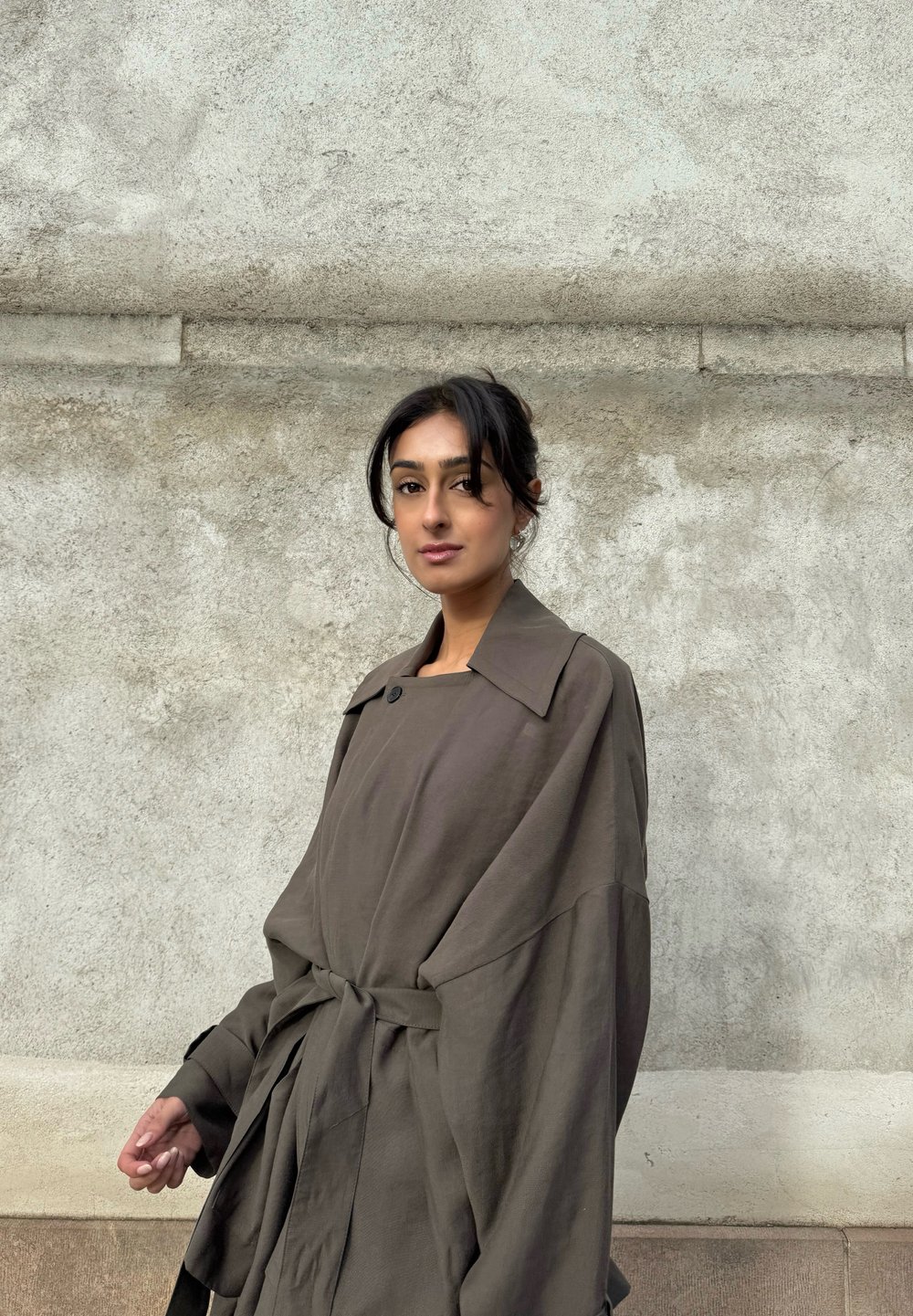 Vrouw met donker haar in een losse knot, gekleed in een oversized bruine jas met een riem, staand tegen een grijze betonnen muur met structuur.