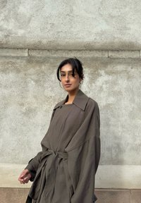 Vrouw met donker haar in een losse knot, gekleed in een oversized bruine jas met een riem, staand tegen een grijze betonnen muur met structuur.