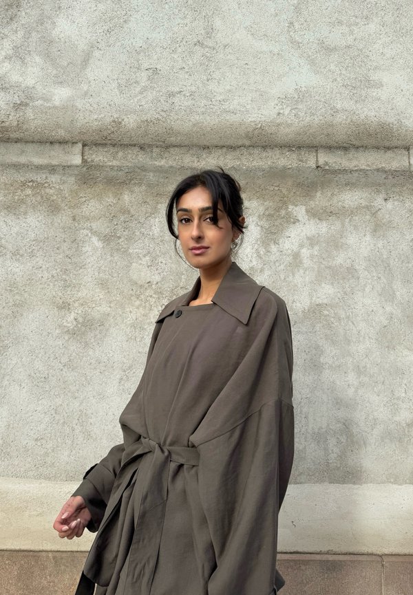 Kvinne med mørkt hår i en løs knute som har på seg en oversized brun frakk med belte, stående mot en teksturert grå betongvegg.