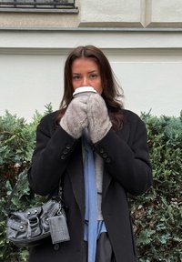 Črn plašč, sivi rokavice iz umetnega krzna, modra karo srajca, drži skodelico kave, črna torbica z reliefnim vzorcem, ozadje z zelenim listjem.