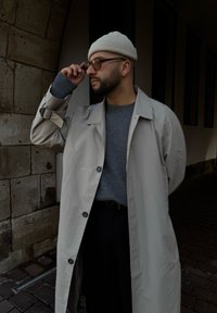 Μπεζ παλτό σε στυλ trench πάνω από γκρι πουλόβερ, συνδυασμένο με ανοιχτό σκουφάκι και γυαλιά. Η υφή είναι λεία, με μινιμαλισικούς μηχανισμούς κουμπώματος και λεπτομέρειες ραφής.