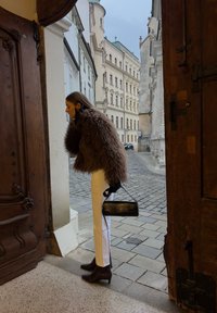 Femme en veste en fourrure marron et pantalon blanc tenant un sac à main noir, debout dans une porte en bois ouverte sur une rue pavée avec des bâtiments historiques.