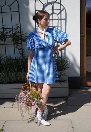 Vestido de cuadros azul y blanco con mangas abullonadas, cintura ajustada; sosteniendo una cesta de mimbre con flores moradas; botas blancas; entorno al aire libre.