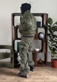 Veste bomber vert olive avec poignets élastiques, pantalon cargo assorti, et bottines noires à talons métalliques, dans un intérieur moderne.
