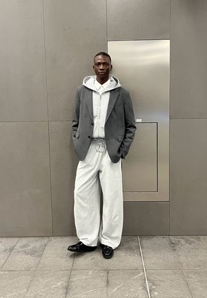Blazer de cuadro gris sobre una sudadera con cremallera gris claro, combinado con pantalones de chándal sueltos grises y zapatos negros. Fondo de una pared gris suave.
