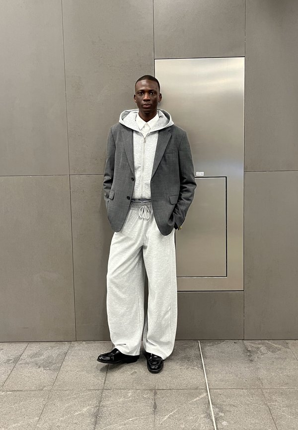 Grijze geruite blazer over een lichtgrijze zip-up hoodie, gecombineerd met losse grijze sweatpants en zwarte schoenen. Achtergrond van een gladde grijze muur.