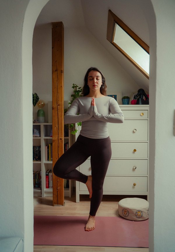 Femme debout sur une jambe en posture de l'arbre de yoga, les mains pressées l'une contre l'autre, les yeux fermés, sur un tapis rose dans une pièce mansardée confortable.