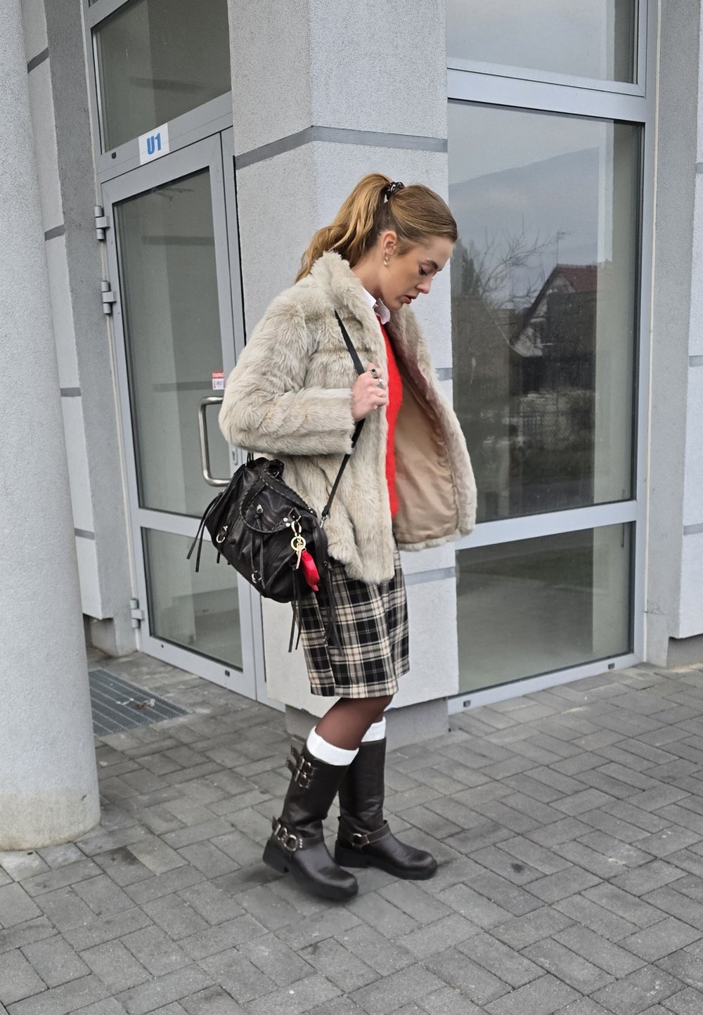 Beige Kunstpelzjacke, roter Pullover, karierter Rock, knielange schwarze Stiefel mit Schnallen und eine schwarze Umhängetasche mit Quaste.