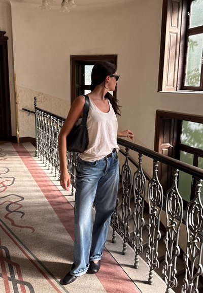 Camiseta sin mangas blanca, jeans azules holgados, zapatos planos negros y un bolso negro; de pie sobre un suelo con patrón junto a una barandilla ornamentada y ventanas.