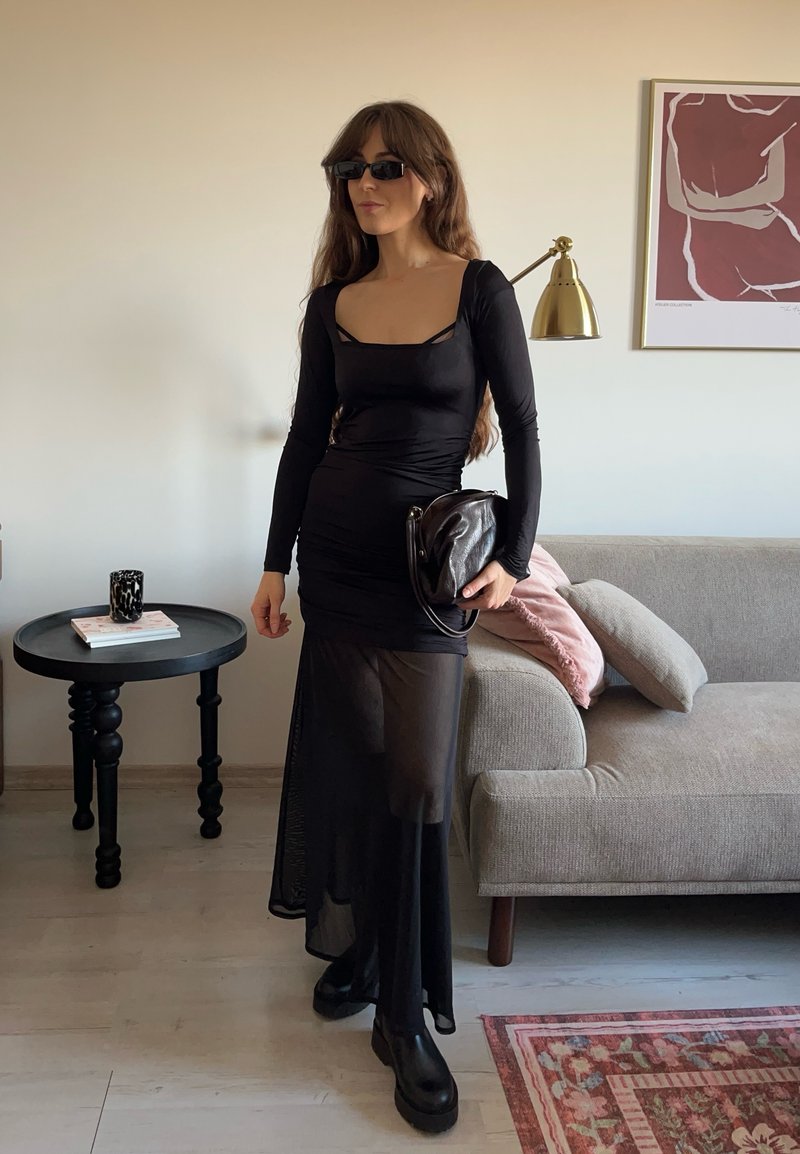 Abito nero a maniche lunghe con scollo quadrato, texture arricciata, gonna trasparente, abbinato a stivali neri massicci e borsa nera.