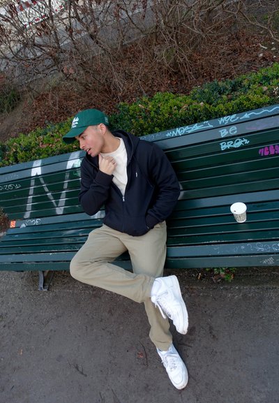 Sudadera con cremallera negra, camisa beige clara, pantalones caqui, zapatillas blancas. Gorra verde sobre cabello corto. Taza de café en un banco verde con grafitis.