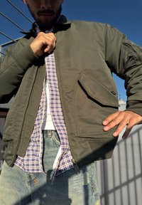 Chaqueta bomber de color verde oliva con cierre de cremallera y puños de canalé, llevada sobre una camisa de cuadros y jeans azules desgastados, que muestra un bolsillo lateral.