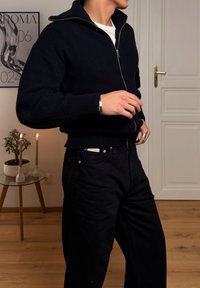 Pull en maille côtelée bleu marine avec fermeture éclair et col montant, accompagné d'un pantalon noir. Les bijoux comprennent un bracelet manchette en argent et des bagues.