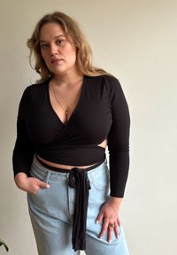 Juodas ilgarankovės apvyniojamos viršus su V formos iškirpte ir šoninėmis išpjovomis, derinamas su šviesiai mėlynais aukštu liemeniu džinsais su dirželiu.